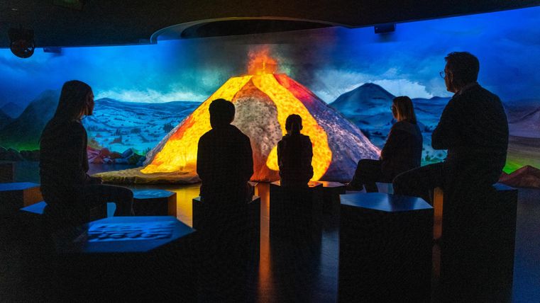 Eine Gruppe von Menschen sitzt vor einer beeindruckenden Vulkanprojektion. Die Szene zeigt einen eruptierenden Vulkan mit lebendigen Farben und einer dramatischen Landschaft.