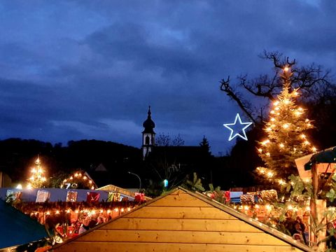 Ein festlich geschmückter Weihnachtsmarkt mit Lichtern und einem geschmückten Baum. Im Hintergrund ist eine Kirche zu sehen, umgeben von einem dunklen, wolkigen Himmel.