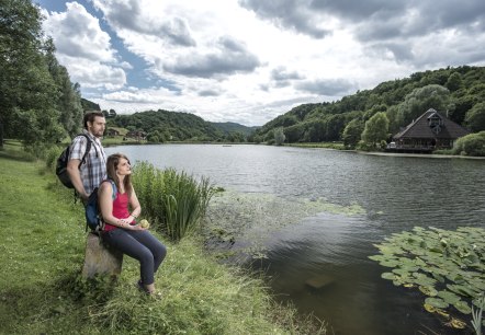 Zwei Personen sitzen am Ufer eines Sees, umgeben von grünen Hügeln und Bäumen. Ein Haus steht am rechten Ufer. Der Himmel ist bewölkt., © Kappest/REMET