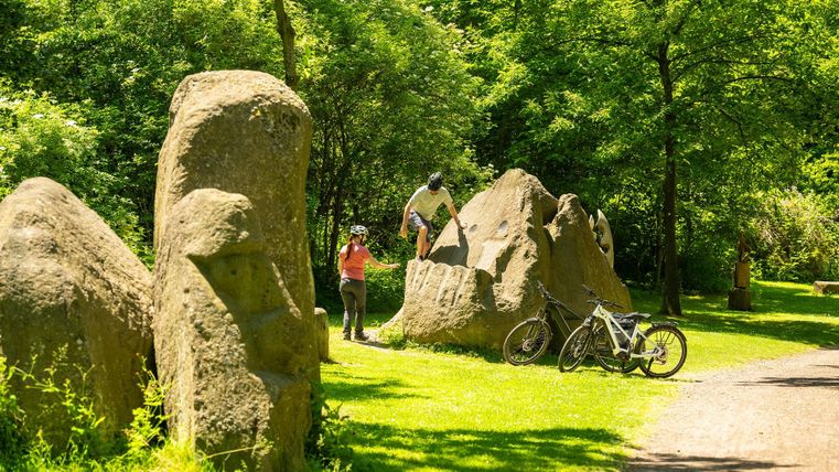 Eine grüne Parklandschaft mit großen Felsen und Radfahrern. Im Hintergrund sind zwei Personen, die die Felsen erkunden.