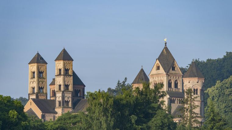 A historic church with several towers stands among trees. The sky is clear and the background features gentle hills.
