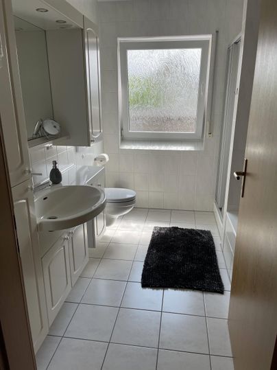 Ein helles Badezimmer mit weißen Fliesen und modernen Sanitärinstallationen. Es gibt ein Fenster, das Tageslicht hereinlässt, und einen schwarzen Teppich auf dem Boden.