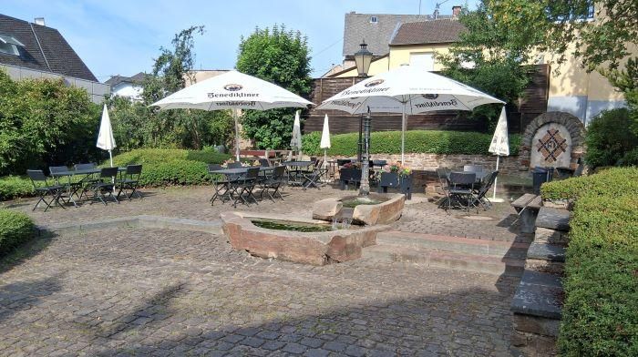Een gezellige buitenruimte met tafels en stoelen onder parasols. Omgeven door groene planten en een schilderachtige stenen façade.