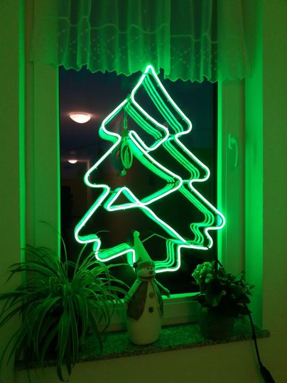 Ein leuchtender grüner Weihnachtsbaum aus Neonröhren hängt an einem Fenster. Neben dem Baum steht eine winterliche Dekoration mit einer Schneemannfigur.