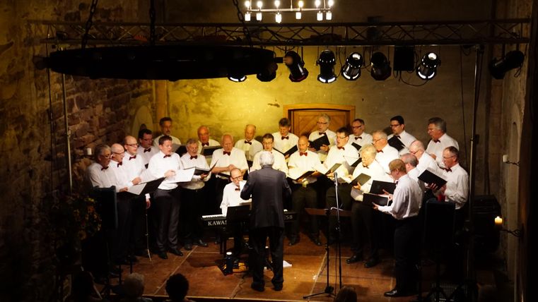 Ein Männerchor in einem historischen Raum singt unter der Leitung eines Dirigenten. Die Sänger tragen weiße Hemden und stehen in mehreren Reihen.