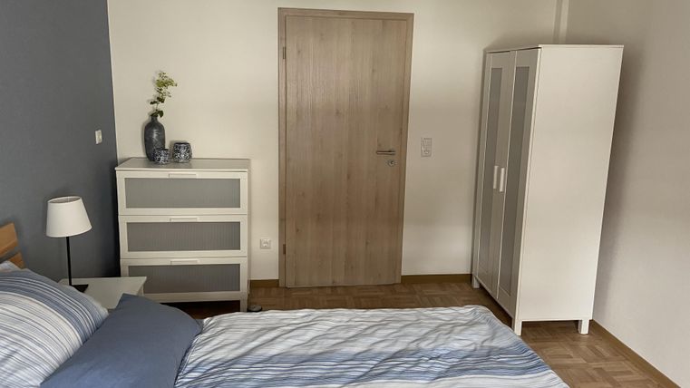 Ein einfaches Schlafzimmer mit einem Bett, einer Kommode und einem Kleiderschrank. Die Wände sind hell gehalten und es gibt eine Lampe an der Decke.