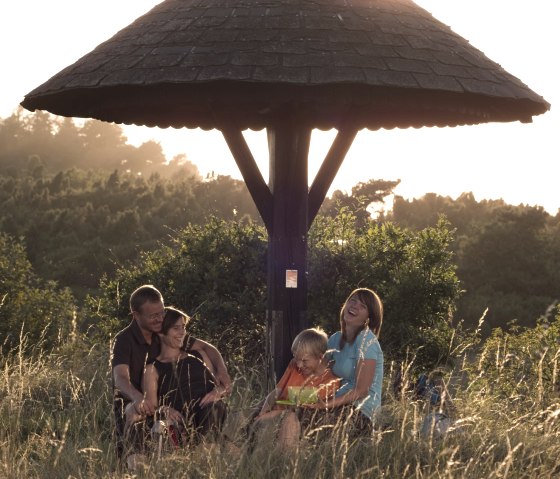 Eine Familie sitzt lachend unter einem hölzernen Unterstand in einer Wiese bei Sonnenuntergang. Im Hintergrund sind Bäume zu sehen., © Traumpfade/Kappest