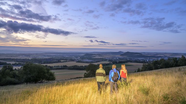 Drei Wanderer stehen auf einem Hügel und betrachten die Landschaft im Abendlicht. Der Himmel ist mit Wolken und warmen Farben gefüllt.