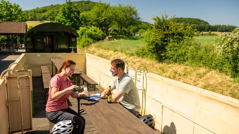A couple is sitting at a table outdoors, enjoying a meal. In the background, green landscapes and an old train car can be seen.