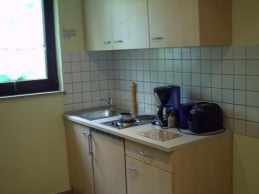 A small, bright kitchen with wooden cabinets and a white countertop. On the countertop, there are various kitchen appliances such as a kettle and a coffee machine.
