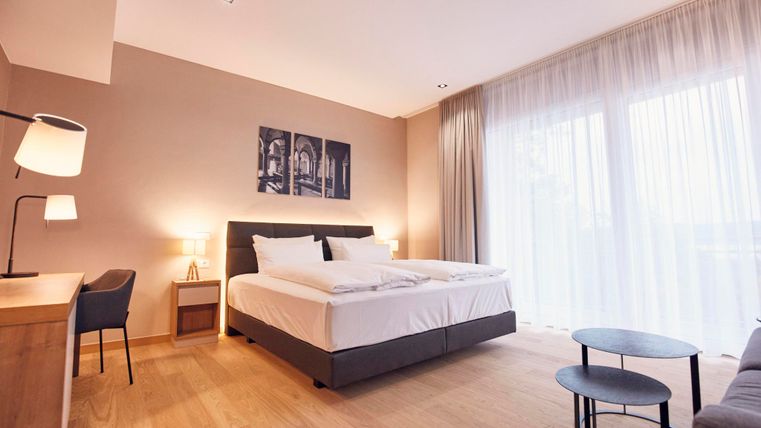 Ein modernes Hotelzimmer mit einem großen Doppelbett und einem Holzfußboden. Das Zimmer ist hell und freundlich mit großen Fenstern und stilvollen Möbeln.