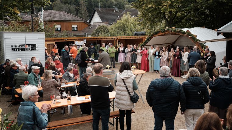 Een feestelijk evenement in de open lucht met veel mensen. In het midden staan groepjes mensen in traditionele klederdracht en er staan banken en tafels voor de gasten.