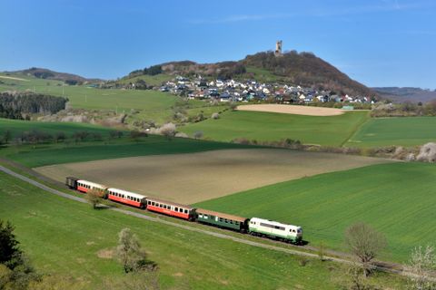 Ein malerischer Zug fährt durch grüne Felder, umgeben von Hügeln. Im Hintergrund ist ein kleiner Ort mit einem Berg und einer Burgruine zu sehen.