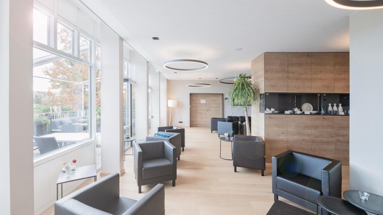 Ein eleganter Empfangsbereich mit modernen Sitzgelegenheiten und großen Fenstern. Die warme Holzgestaltung und Pflanzen schaffen eine einladende Atmosphäre.