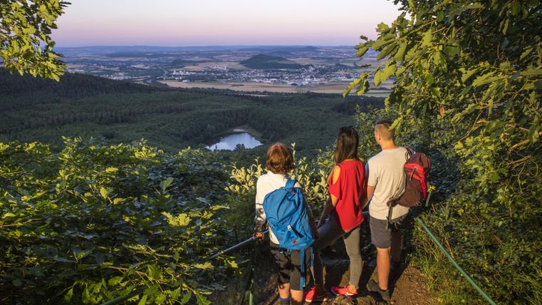 Drie wandelaars genieten van het uitzicht op een schilderachtig landschap met bossen en een meer. De zon gaat langzaam onder en dompelt het tafereel in warm licht.