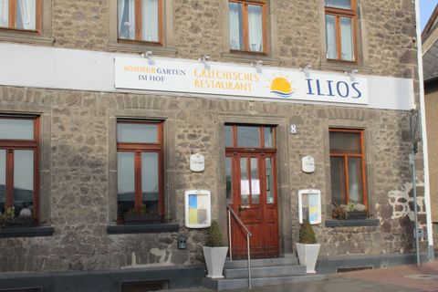 Das Bild zeigt die Fassade eines griechischen Restaurants namens "Ilios". Der Eingang ist einladend mit Fenster und Blumenkübeln.