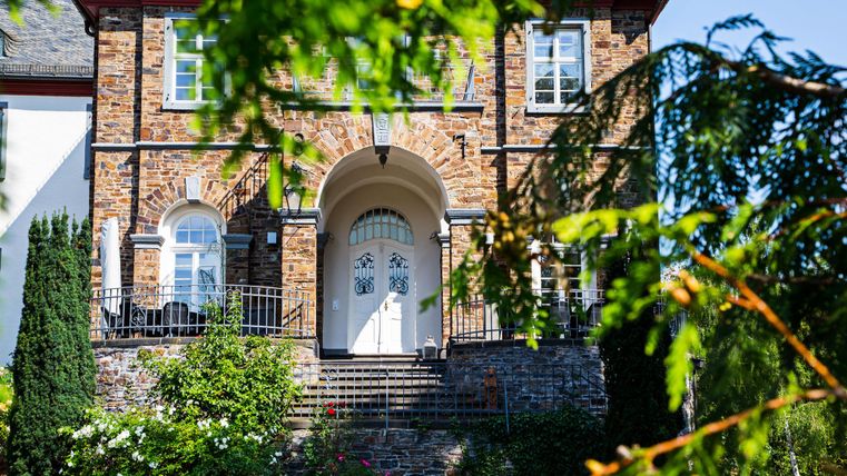 A beautiful house with a historic facade made of stones and a white front door.
Surrounded by green plants and a clear blue sky.