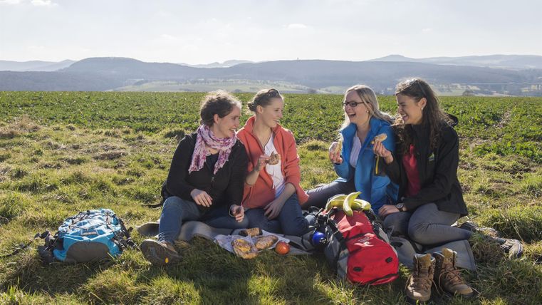 Vier Frauen sitzen auf einer Wiese und genießen ein Picknick. Im Hintergrund erstreckt sich eine malerische Landschaft.