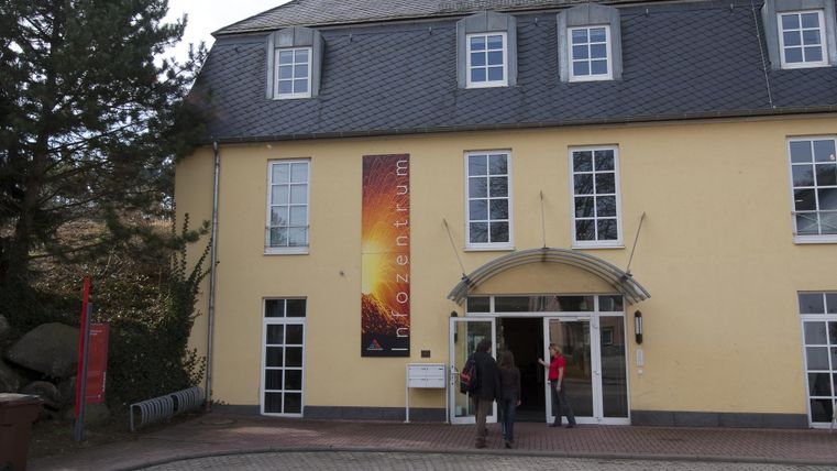 A modern building with a yellow facade and large windows. Two people are standing in front of the entrance.