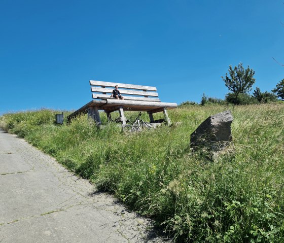 Eine riesige Holzbank steht auf einer gr&uuml;nen Wiese unter klarem Himmel. Eine Person sitzt darauf, daneben ein Fahrrad. Ein Weg f&uuml;hrt zur Bank., &copy; Svenja Schulze-Entrup