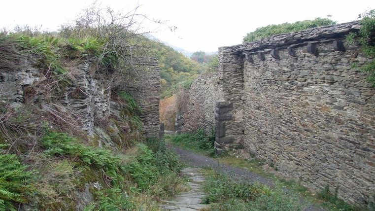 A narrow path leads through an old stone wall, surrounded by green vegetation. The landscape appears calm and deserted.