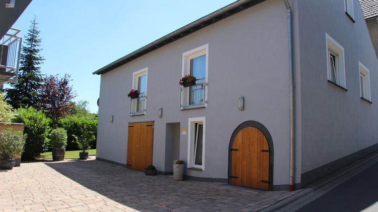 Ein hellgraues Haus mit Holzfenstern und Türen. Der Eingangsbereich ist gepflastert und von Pflanzen umgeben.