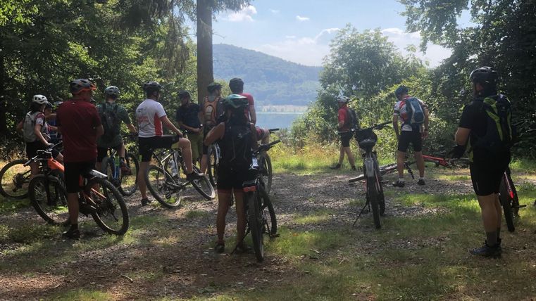 Eine Gruppe von Radfahrern steht im Wald und genießt die Aussicht auf einen See. Die Umgebung ist grün und sonnig.
