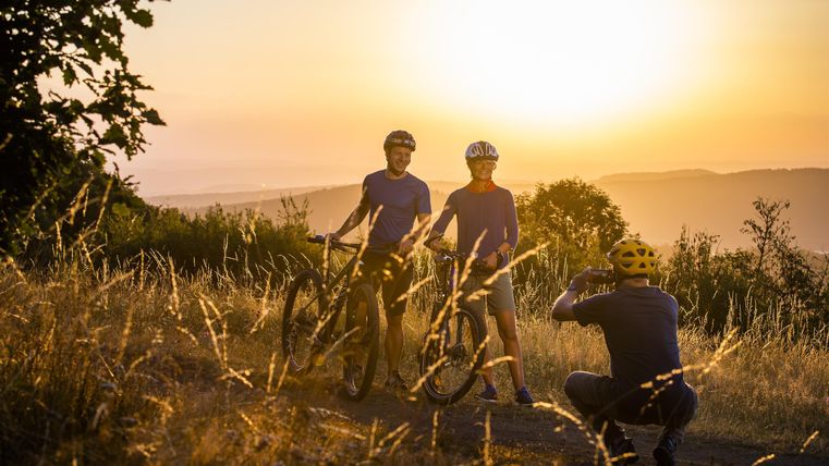 Zwei Radfahrer stehen in der Natur bei Sonnenuntergang. Ein Fotograf hält die Szene fest und fängt die goldene Lichtstimmung ein.
