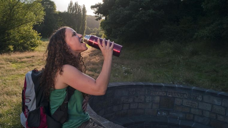 Eine wandernde Person trinkt aus einer Thermosflasche. Im Hintergrund sind Bäume und eine grüne Wiese zu sehen.
