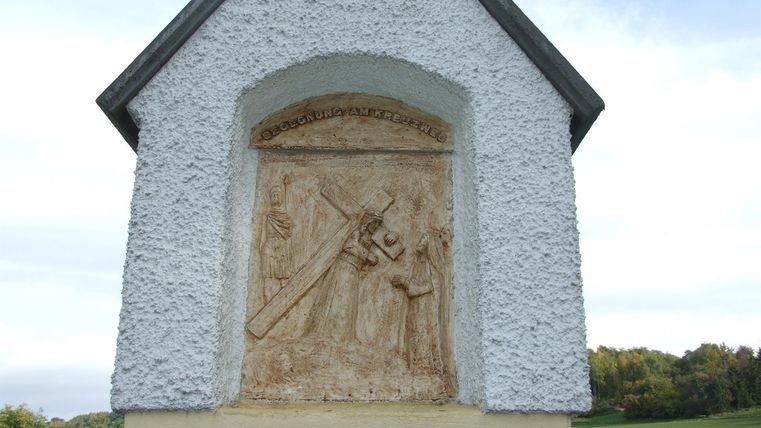 Eine Schnitzerei in einem weißen Nischengebäude, die zwei Figuren zeigt. Der Hintergrund besteht aus einer ländlichen Landschaft.