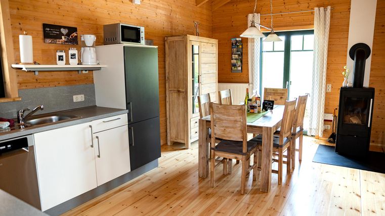 A cozy interior view of a wooden apartment with a modern kitchenette and a dining area. The large table is surrounded by wooden chairs, and the window provides plenty of daylight.