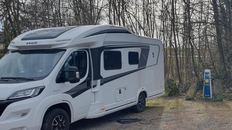 A white motorhome is parked in a spot in the woods. In the background, you can see a charging station for electric vehicles.