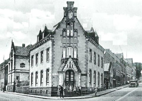 Ein historisches Gebäude mit markanten architektonischen Details und einem Uhrenturm. Die Straße führt entlang der Fassade, und einige Personen stehen davor.