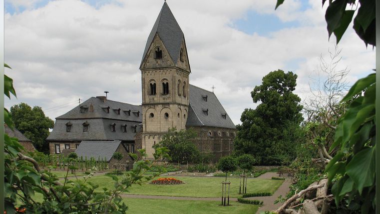 Eine historische Gebäude mit einem hohen Turm, umgeben von einem gepflegten Garten. Der Himmel ist teilweise bewölkt und die Atmosphäre ist ruhig.