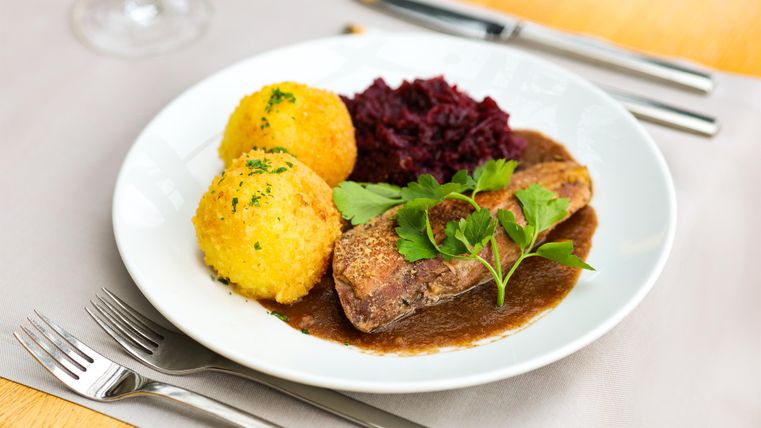 Ein Teller mit zartem Fleisch, serviert mit Knödeln und Rotkohl. Das Gericht ist mit frischen Kräutern garniert.