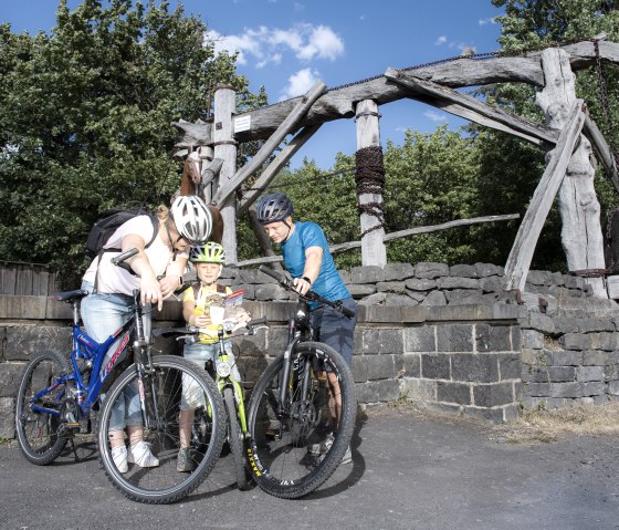 Eine Familie mit Fahrr&auml;dern betrachtet eine Karte vor einem historischen Bergbauger&auml;t aus Holz. Der Himmel ist blau und die Umgebung gr&uuml;n., &copy; Klaus-Peter Kappest/Vulkanregion Laacher See