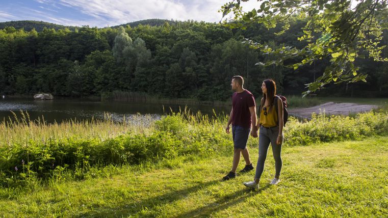 A couple is walking along a calm body of water in a green landscape. In the background, gentle hills and a blue sky can be seen.