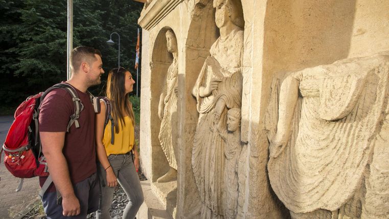 Ein Paar betrachtet eine sculpturale Wand mit historischen Figuren. Die Atmosphäre ist entspannt und die Umgebung ist von natürlicher Schönheit geprägt.