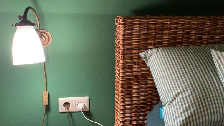 Ein stilvolles Bett mit einem geflochtenen Kopfteil und gestreiften Kissen. Neben dem Bett hängt eine moderne Lampe an der wandgrünen Wand.