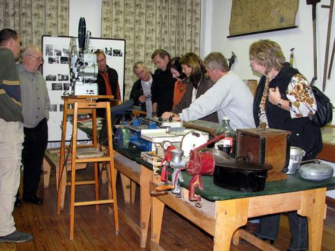 Eine Gruppe von Menschen betrachtet historische Werkzeuge und Geräte auf einem Tisch. Im Hintergrund sind Fotos und alte Karten zu sehen.