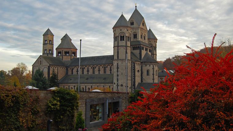 Eine beeindruckende Kirche mit mehreren Türmen steht vor einem bewölkten Himmel. Im Vordergrund leuchten rote Herbstblätter.