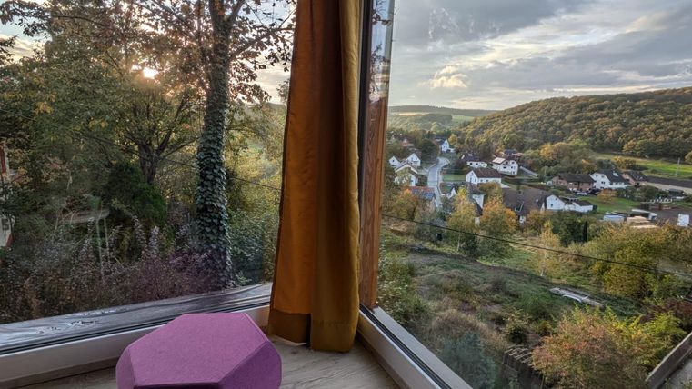 Ein gemütlicher Blick aus einem Fenster auf eine malerische Landschaft. Im Vordergrund steht ein lila Hocker, während die Sonne hinter den Bäumen scheint.