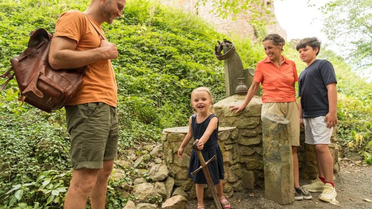 Eine Familie steht an einer alten Steinsäule im Freien. Ein kleines Mädchen hält ein Schwert und lächelt, während die anderen Familienmitglieder aufmerksam zuschauen.
