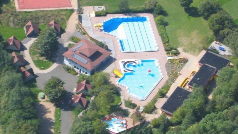 Ein Luftbild eines Freizeitgeländes mit mehreren Schwimmbecken und einer Unterkunft. Umgeben von Bäumen und Freiflächen für Aktivitäten.