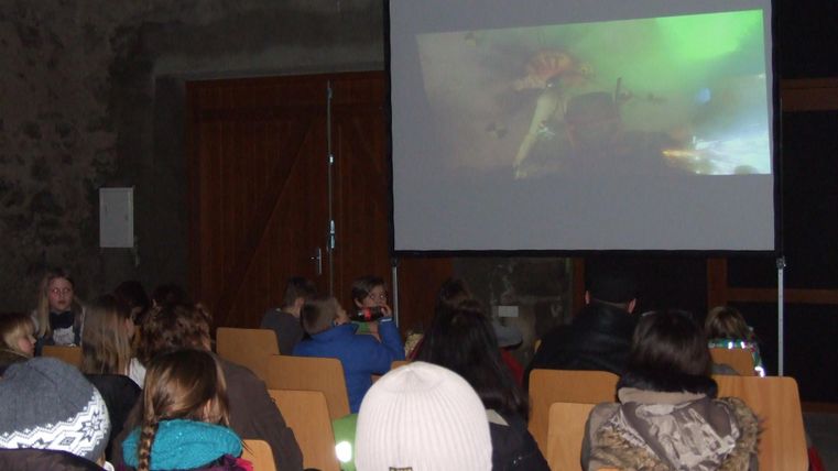 A group of people is sitting in a room watching a movie on a large screen. The room has a rustic atmosphere with stones and wood.