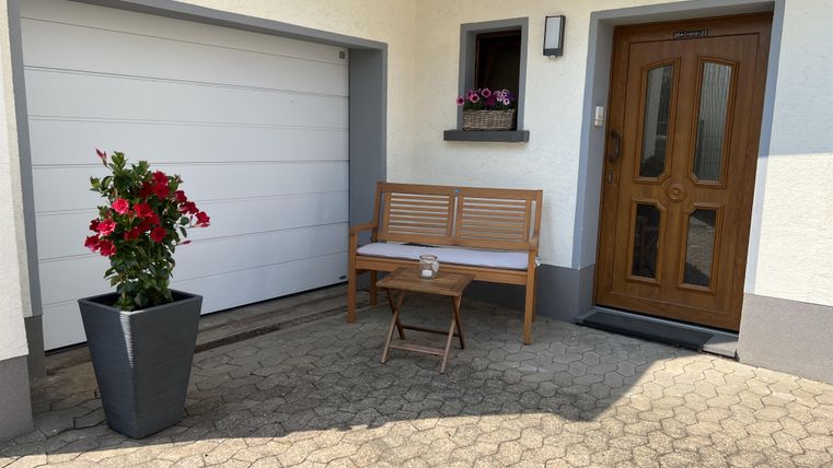 Ein einladender Eingangsbereich mit einer Holzbank und blühenden Pflanzen. Zu sehen sind auch eine Garage und eine Haustür aus Holz.