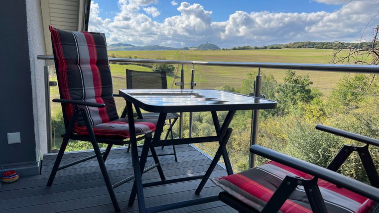 A cozy balcony with a table and two chairs. In the background, green fields and a blue sky can be seen.