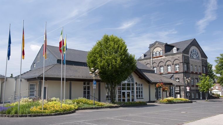 A modern building with flags in the foreground and a group of green trees. The sky is clear and blue.