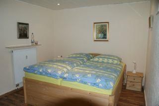Ein einfaches Schlafzimmer mit einem Doppelbett und bunten Bettdecken. An der Wand hängen zwei Bilder und ein Heizkörper ist sichtbar.