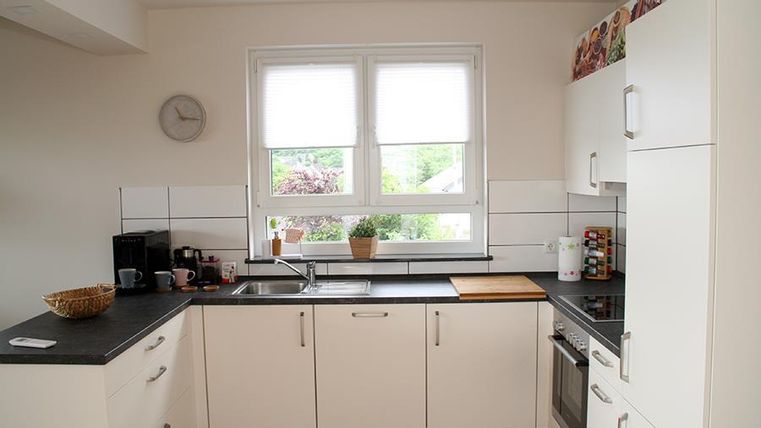 Eine moderne Küche mit weißen Schränken und einem schwarzen Arbeitsbereich. Große Fenster sorgen für viel Tageslicht und eine freundliche Atmosphäre.